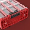 Czerwony organizer z przezroczystą pokrywą, podzielony na kilka przegródek, z widocznym napisem "QBRICK SYSTEM" na boku. Pokrywa jest zabezpieczona czarnym zatrzaskiem.
