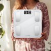 Osoba trzyma białą wagę łazienkową z wyświetlaczem pokazującym "63.0 kg" oraz logo "ALPHA" na dole urządzenia. W tle widoczne są rośliny i zasłony, a osoba ubrana jest w wzorzystą bluzkę.