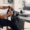 Osoba siedzi przy biurku, trzymając kierownicę symulatora wyścigowego z zamontowanym kontrolerem LOGITECH RS Shifter & Handbrake. Na biurku znajduje się monitor z wyświetloną grą wyścigową oraz głośnik i myszka.