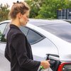 Na zdjęciu widać osobę podłączającą ładowarkę do samochodu elektrycznego, stojącego na tle zieleni. Samochód jest koloru srebrnego, a ładowarka ma czarny uchwyt z zielonym akcentem.