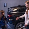 Na zdjęciu widać czarny samochód elektryczny zaparkowany w garażu, obok którego stoi ładowarka ścienna Green Cell HabuDen Wallbox EVGC021A2275 22kW. Dwie osoby trzymają kabel do ładowania, przygotowując się do podłączenia go do samochodu.