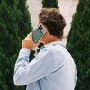 Na zdjęciu widoczna jest osoba trzymająca przy uchu telefon z przymocowanym powerbankiem. W tle znajdują się zielone drzewa i fragment jasnego budynku.