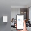 Na zdjęciu widać nowoczesne wnętrze salonu z białym włącznikiem światła zamontowanym na ścianie oraz rękę trzymającą smartfon z aplikacją do sterowania oświetleniem, na ekranie widoczne są trzy przyciski z ikoną włączania i podpisami "Light1".