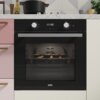 Czarny piekarnik elektryczny z wyświetlaczem pokazującym godzinę 11:40, umieszczony w zabudowie kuchennej z marmurowym blatem, na którym stoją różne akcesoria kuchenne. Na drzwiczkach piekarnika widoczne jest logo "FRANKE".