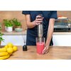 Osoba trzyma blender ręczny w kolorze srebrno-czarnym, miksując różowy płyn w przezroczystym naczyniu na drewnianym blacie. Obok znajdują się cytryny, banany, trzepaczka oraz rośliny w doniczkach, a w tle widać piekarnik.