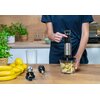 Na zdjęciu znajduje się blender ręczny w kolorze srebrno-czarnym, trzymany nad pojemnikiem z pokrojonymi owocami. Obok widoczne są akcesoria do blendera, banany, cytryny oraz doniczki z ziołami.