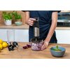 Osoba trzyma blender ręczny GÖTZE & JENSEN HB902X w kolorze srebrno-czarnym, który znajduje się w pojemniku z posiekaną cebulą. Na blacie widoczne są akcesoria do blendera, połówki cebuli, cytryny oraz miska z potrawą udekorowaną listkami mięty.