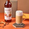 Na zdjęciu znajduje się butelka syropu z etykietą "Le Sirop de MONIN Pumpkin Spice" oraz szklanka z napojem, na której widać bitą śmietanę i posypkę. Obok leżą przyprawy, takie jak laska cynamonu i goździki, na pomarańczowym tle.