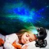 Na zdjęciu widać projektor w kształcie czarnego astronauty, który wyświetla kolorowe światła na tle przypominającym gwiaździste niebo. Obok projektora leży dziecko z zamkniętymi oczami, przytulające pluszowego misia.