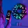 Na zdjęciu widać osobę trzymającą smartfon z aplikacją wyświetlającą kolorowe kropki w kształcie choinki, a w tle znajduje się świecący wieniec świąteczny z kolorowymi światłami. Na ekranie telefonu widoczny jest napis "Pole Rainbow".