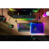 Na zdjęciu znajduje się biurko z klawiaturą o podświetleniu RGB, myszką na podkładce również z podświetleniem oraz słuchawkami i mikrofonem. Na monitorze widoczne jest logo Razer, a na biurku znajduje się także głośnik i kolorowa lampa.