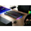 Na zdjęciu znajduje się klawiatura z podświetleniem RGB na drewnianym biurku, obok podkładki z myszką i kolorowego głośnika. Klawiatura ma ciemną obudowę i podświetlane klawisze, a w tle widoczne są elementy stanowiska komputerowego.