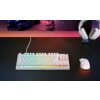 Na zdjęciu znajduje się biała klawiatura z podświetleniem RGB, myszka z perforowaną obudową oraz słuchawki na stojaku, umieszczone na ciemnym biurku. Na klawiaturze widoczny jest napis "SteelSeries".