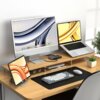 Na zdjęciu znajduje się biurko z drewnianym blatem, na którym ustawiono monitor, laptop, tablet oraz klawiaturę i myszkę na czarnej podkładce. Obok monitora znajduje się mała roślina w doniczce, a pod półką leżą okulary.