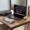 Na zdjęciu znajduje się laptop z ciemnym ekranem i ikonami aplikacji na dolnym pasku, obok którego leży klawiatura o szarych i białych klawiszach oraz filiżanka kawy z mleczną pianką. W tle widać szarą torbę na laptopa i żaluzje na oknie.