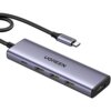 Na zdjęciu widoczny jest hub USB w kolorze srebrnym z czterema portami USB i jednym portem USB typu C, z napisem "UGREEN" na obudowie. Kabel z wtyczką USB typu C również ma napis "UGREEN".