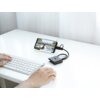 Na zdjęciu widać biały stół, na którym znajduje się klawiatura oraz smartfon w uchwycie, podłączony do czarnego huba USB. Na ekranie smartfona wyświetlany jest krajobraz, a hub ma widoczne logo producenta.
