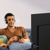 Mężczyzna w słuchawkach siedzi na kanapie, obok niego leży osoba z zamkniętymi oczami. Na telewizorze podłączony jest adapter Bluetooth UGREEN.