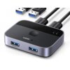 Na zdjęciu widoczny jest przełącznik UGREEN z dwoma portami USB oznaczonymi jako "OUTPUT 1" i "OUTPUT 2", oraz przyciskiem na górze obudowy. Obok urządzenia znajdują się dwa kable USB z logo UGREEN na wtyczkach.
