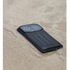 Na zdjęciu widoczny jest szary smartfon z wyraźnie zaznaczonymi elementami aparatu na tylnej obudowie, leżący na powierzchni przypominającej piasek lub beton. Obudowa telefonu ma teksturowaną powierzchnię i widoczny czerwony przycisk z boku.