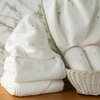Na zdjęciu widoczne są białe ręczniki z delikatnymi kolorowymi kropkami, ułożone w stos oraz w wiklinowym koszu na drewnianej powierzchni. W tle znajduje się marmurowa ściana, a ręczniki mają różnokolorowe lamówki.
