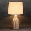 Beżowa lampa stołowa z abażurem, posiadająca ozdobną podstawę z wzorem w jodełkę, stoi na tle ciemnej, teksturowanej ściany.