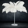 Lampa podłogowa z białymi piórami na metalowej podstawie, ustawiona na ciemnym tle. Produkt ma elegancki i dekoracyjny wygląd.
