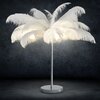 Na zdjęciu znajduje się lampa podłogowa z białymi piórami, które otaczają źródła światła, na tle ciemnej, teksturowanej ściany. Lampa ma metalową podstawę i prosty, cylindryczny trzon.