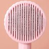 Różowa szczotka do sierści z metalowymi igłami, zakończonymi białymi kuleczkami, na tle w kolorze brzoskwiniowym. Szczotka ma okrągły kształt z falistym brzegiem.