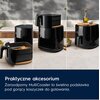 Na zdjęciu widoczna jest czarna frytkownica beztłuszczowa z logo Electrolux, umieszczona na blacie kuchennym, z drewnianą podstawką i koszyczkiem z frytkami. Na dole zdjęcia znajduje się tekst: "Praktyczne akcesorium Żaroodporny MultiCoaster to świetna po