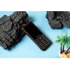 Telefon komórkowy z czarną obudową i pomarańczowymi akcentami leży na niebieskim tle, otoczony sztucznymi skałami i miniaturową palmą. Na ekranie telefonu widoczne jest logo "ulefone", a poniżej znajduje się klawiatura numeryczna.