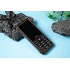 Na zdjęciu znajduje się czarny telefon z pomarańczowymi akcentami, leżący na niebieskim tle obok ciemnych, teksturowanych kamieni i zielonych liści. Na ekranie telefonu widoczne jest logo "ulefone", a poniżej znajduje się klawiatura numeryczna z przyciska