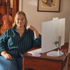 Na zdjęciu znajduje się drewniane biurko, na którym stoi komputer typu All-in-One z klawiaturą i myszką. Na ekranie komputera widoczny jest interfejs wideokonferencji z kilkoma osobami.