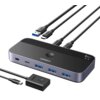 Na zdjęciu znajduje się przełącznik KVM marki UGREEN z kilkoma portami USB i kablami, z widocznym napisem "UGREEN" oraz oznaczeniem "5Gbps". Produkt ma elegancki, nowoczesny design z czarną i srebrną kolorystyką.