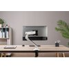 Na zdjęciu znajduje się biurko z zamontowanym uchwytem do monitora marki ULTRADESK, na którym umieszczony jest monitor. W tle widoczna jest szara ściana, regał z książkami oraz roślina doniczkowa.