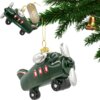 Zielona bombka w kształcie samolotu z czerwonymi oknami i srebrnymi elementami, zawieszona na złotym sznurku, obok gałązki choinki. Na etykiecie widnieje napis "CHRISTMAS DECORATION".