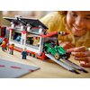 Na zdjęciu znajduje się zestaw klocków LEGO przedstawiający ciężarówkę z bolidami wyścigowymi, z widocznymi szczegółami konstrukcji, takimi jak rampy i detale pojazdów. Obok ciężarówki znajdują się figurki LEGO, a w tle widać uśmiechniętą osobę, która baw