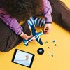 Na zdjęciu widać osobę składającą model samolotu z klocków LEGO, siedzącą na żółtym tle. Obok znajduje się tablet z wyświetlonym schematem oraz kilka luźnych elementów LEGO.