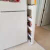 Białe, wąskie półki regału wypełnione są różnorodnymi słoikami i butelkami, umieszczone obok białej lodówki. W tle widoczne są drzwi oraz fragmenty podłogi wyłożonej wykładziną i płytkami.