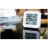 Na zdjęciu znajduje się biały czujnik temperatury i wilgotności z wyświetlaczem pokazującym godzinę 15:07, temperaturę 26.0°C oraz wilgotność 39%. Urządzenie stoi na błyszczącej powierzchni, na której odbija się jego obraz.