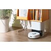 Na zdjęciu znajduje się robot sprzątający SWITCHBOT K10+ na podłodze obok stacji dokującej, która jest umieszczona pod drewnianym regałem z książkami. Obok regału stoi duża donica z rośliną.