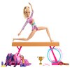 Lalka Barbie Gimnastyczka HRG52 wykonuje ćwiczenie na brązowej równoważni, ubrana w kolorowy strój gimnastyczny. Wokół znajdują się akcesoria, takie jak złoty puchar, różowe pałeczki, hula-hoop, torba sportowa, butelka na wodę, banan, fioletowe buty i kol