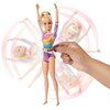 Na zdjęciu znajduje się lalka Barbie w stroju gimnastycznym w kolorach różowym, złotym, niebieskim i fioletowym, z uniesionymi rękami. Obok lalki widoczna jest dłoń, która trzyma mechanizm obracający lalkę, a w tle widać rozmyte sylwetki lalki w ruchu.