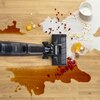 Na drewnianej podłodze rozlane są różne substancje, w tym kawa, mleko z płatkami, jajko oraz owoce, a obok nich znajduje się mop elektryczny z widocznym logo "Tefal". Mop jest w trakcie sprzątania, co sugeruje jego skuteczność w usuwaniu różnorodnych zabr
