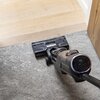 Na obrazie widoczny jest elektryczny mop z wyświetlaczem pokazującym liczbę "99" oraz napisem "TEFAL" na głowicy czyszczącej. Urządzenie znajduje się na granicy dwóch powierzchni: drewnianej podłogi i szarego dywanu.