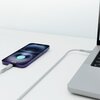 Na zdjęciu widoczny jest szary kabel USB Typ-C - Lightning podłączony do laptopa i smartfona, który pokazuje ikonę ładowania z napisem "100%". Kabel leży na białej powierzchni, a laptop ma widoczną część klawiatury.