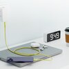 Na zdjęciu znajduje się żółty kabel USB-C - USB-C podłączony do telefonu leżącego na laptopie, obok którego znajdują się słuchawki bezprzewodowe i zegar cyfrowy pokazujący godzinę 9:49. W tle widoczny jest kubek z napojem.