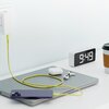 Na zdjęciu znajduje się żółty kabel USB Typ-C - Lightning podłączony do fioletowego telefonu, leżącego na zamkniętym laptopie. Obok widoczny jest zegar elektroniczny z wyświetlaną godziną 9:49, etui na słuchawki, słuchawki douszne oraz kubek z napojem.