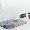 Na zdjęciu znajduje się różowy kabel USB-C - USB-C podłączony do telefonu leżącego na zamkniętym laptopie, obok którego znajdują się słuchawki bezprzewodowe i ich etui. W tle widoczny jest zegar elektroniczny z wyświetlaczem pokazującym godzinę 9:49 oraz 