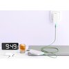 Na zdjęciu widać zielony kabel USB Typ-C - Lightning podłączony do gniazdka i telefonu, który leży na zamkniętym laptopie. Obok znajduje się zegar z wyświetlaczem pokazującym godzinę 9:49, filiżanka z napojem oraz etui na słuchawki bezprzewodowe.